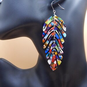 May include: A pair of long, colourful dangle earrings with a leaf design. The earrings are made of metal and have a silver finish. The leaves are layered and have a multi-coloured pattern.