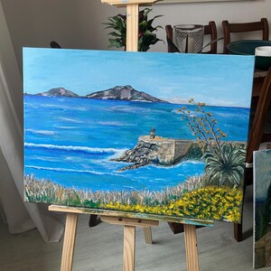 Pode incluir: Pintura a óleo original de uma cena costeira. A obra retrata um oceano azul brilhante com ondas, um cais de pedra e um farol. O primeiro plano apresenta folhagem amarela e verde, com duas ilhas ao fundo. A pintura é exibida em um cavalete de madeira.