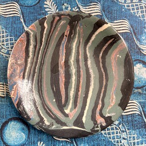 May include: A round ceramic plate with a marbled design in shades of black, green, white, and peach. The plate has a glossy finish and sits on a blue patterned surface. The plate's design resembles a zebra pattern.
