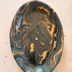 May include: Oval ceramic dish with a marbled pattern in teal, black, gold, and brown. The swirling design has a glossy finish and a slightly raised edge. This decorative dish is suitable for serving or display. The measurements are in centimetres.