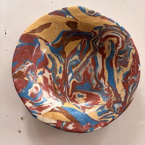 May include: A handmade, round bowl with a marbled design. The bowl features a blend of colors, including red, blue, tan, and brown, creating a unique, swirled pattern. The bowl is likely made of ceramic or a similar material.