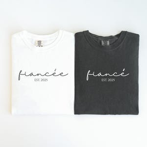May include: Two t-shirts, one white and one dark gray, are displayed. Each shirt has the word "fianc&eacute;e" in cursive script, with "EST. 2025" below it. The shirts are folded and laid flat.