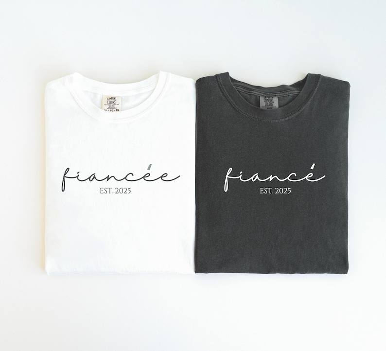 May include: Two t-shirts, one white and one dark gray, are displayed. Each shirt has the word "fianc&eacute;e" in cursive script, with "EST. 2025" below it. The shirts are folded and laid flat.