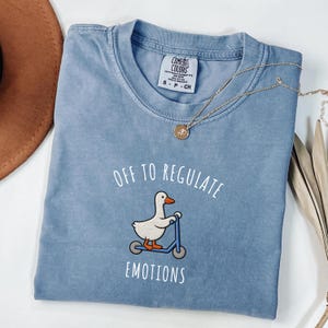 May include: A light blue Comfort Colors t-shirt featuring the text "OFF TO REGULATE EMOTIONS" and a cartoon duck on a blue scooter. A gold necklace with a pendant is on the shirt.