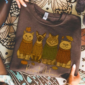 May include: A brown sweatshirt featuring four cats wearing sweaters, with autumn leaves and a pumpkin graphic. The text "Purrfect Autumn" is printed in a gold script. The sweatshirt is held by a person.