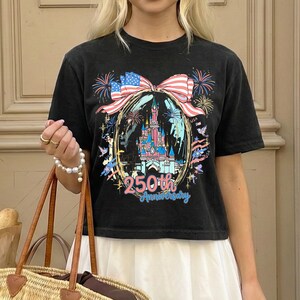 K&ouml;nnte beinhalten: Schwarzes T-Shirt mit einer farbenfrohen Grafik eines Schlosses, Feuerwerks und einer Schleife mit amerikanischer Flagge. Der Text lautet "250th Anniversary" in Gold. Das T-Shirt wird von einer Person getragen.