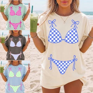 Puede incluir: Una camiseta de manga corta con un estampado gráfico de un bikini de dos piezas con patrón de cuadros. El diseño incluye un top triangular con tiras laterales para anudar y una braguita de bikini a juego en varios colores sólidos.
