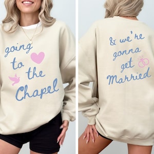 May include: Beige sweatshirt with the text "going to the Chapel" in blue script, a pink heart, and a dove graphic. The back of the sweatshirt says "& we're gonna get Married" in blue script with pink wedding rings.