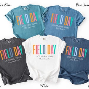 May include: Five t-shirts in shades of blue, grey, and white. Each shirt displays the text "FIELD DAY" in colourful block letters. The school name and a teacher's name are printed below the main text.