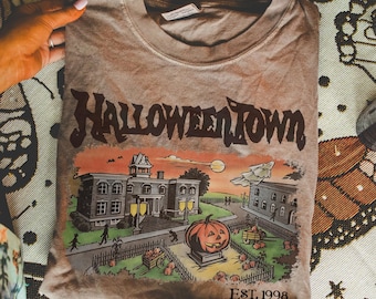 May include: A brown t-shirt with a graphic of a Halloween scene, including a large pumpkin, buildings, and the text "Halloweentown EST. 1998". The shirt is folded and held by hands.