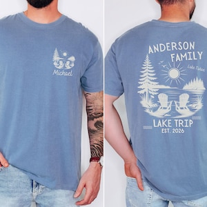 May include: A blue t-shirt featuring a small graphic on the front left chest with a sun, trees, and the name Michael. The back displays a large graphic of two chairs by a lake, trees, a sun, and the text Anderson Family Lake Trip Est. 2026.