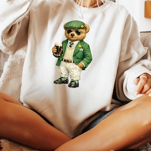 May include: White sweatshirt featuring a cartoon bear wearing sunglasses, a green jacket, and a green hat. The bear is holding a glass of dark beer. The bear is wearing white pants, a white sweater vest, and black shoes with green shamrocks.