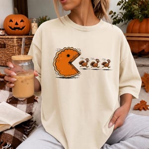 May include: A cream-colored short-sleeve t-shirt featuring a graphic print of an orange pumpkin pie slice shaped like a video game character chasing three brown turkeys. The design is centered on the chest of the garment, which has a crew neckline.