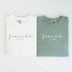 May include: Two folded t-shirts, one white and one sage green, with the word "fianc&eacute;e" in cursive and "EST. 2025" printed below. The shirts are made of a soft material and are likely for a couple.