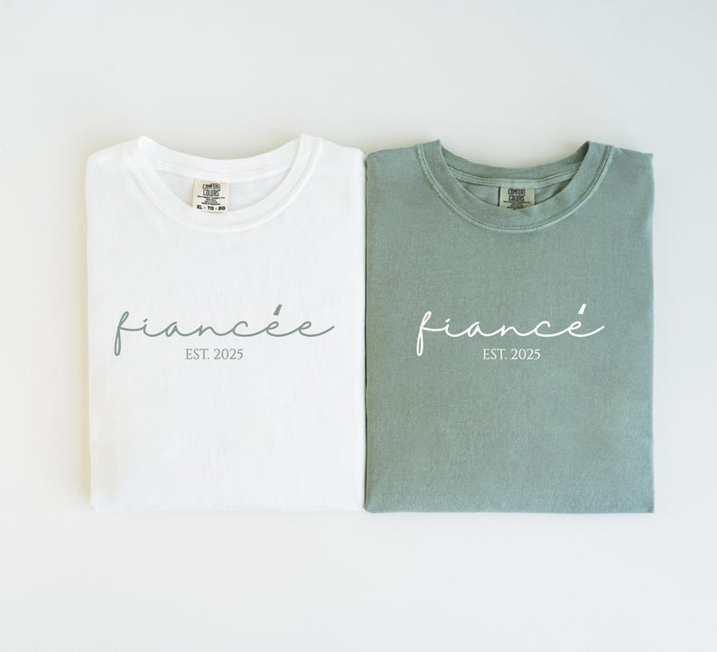 May include: Two folded t-shirts, one white and one sage green, with the word "fianc&eacute;e" in cursive and "EST. 2025" printed below. The shirts are made of a soft material and are likely for a couple.