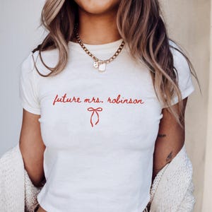 May include: White t-shirt with the red text "future mrs. robinson" and a red bow graphic. The shirt has a crew neck and short sleeves. A gold chain necklace with charms is visible.