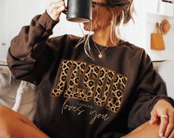 Op de afbeelding: Een bruine sweatshirt met het woord "JESUS" in een luipaardprint. Onder het woord "JESUS" staat de zin "loves you" en de tekst "1 JOHN 4:19" in een handschrift.