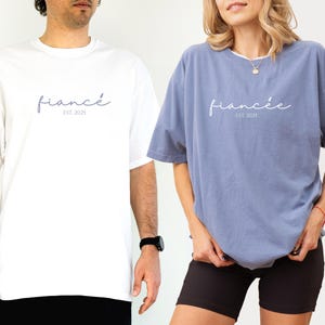 May include: White and blue t-shirts with the word "fianc&eacute;" and "fianc&eacute;e" printed in a script font. The text "EST. 2025" is below the words. The white shirt is worn by a person on the left, and the blue shirt is worn by a person on the right.