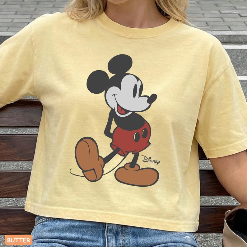 May include: Butter yellow t-shirt featuring a classic Mickey Mouse graphic. The cartoon character is depicted in black, red, brown, and white, with the Disney logo. The shirt has short sleeves and a relaxed fit.