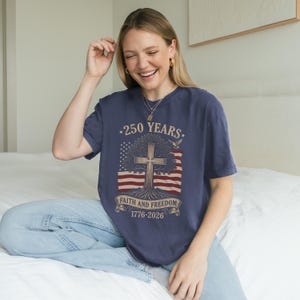 May include: Navy blue t-shirt with a graphic commemorating 250 years. The design features a cross within a tree, the American flag, and the text "Faith and Freedom 1776-2026".