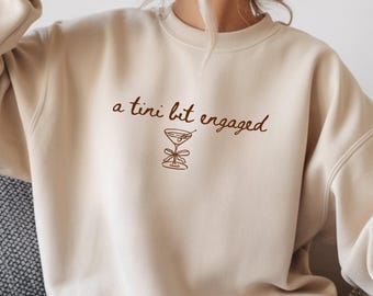 May include: Beige sweatshirt with the phrase "a tini bit engaged" in brown script. A brown line drawing of a martini glass with a bow is below the text. The sweatshirt has a crew neck and long sleeves.