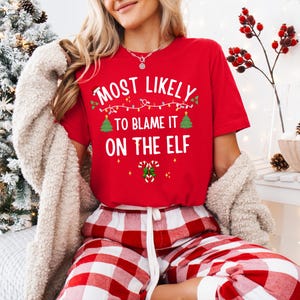 May include: A red short-sleeved t-shirt featuring the white text "MOST LIKELY TO BLAME IT ON THE ELF" with graphic illustrations of a string of Christmas lights, two small green pine trees, and a pair of candy canes with a green bow.