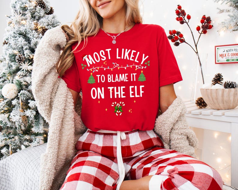 May include: A red short-sleeved t-shirt featuring the white text "MOST LIKELY TO BLAME IT ON THE ELF" with graphic illustrations of a string of Christmas lights, two small green pine trees, and a pair of candy canes with a green bow.