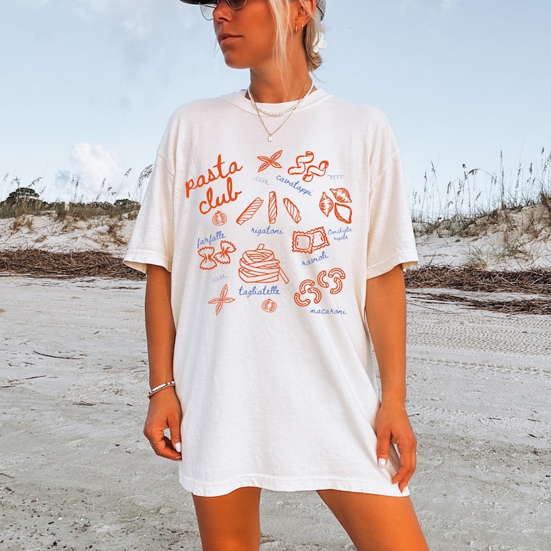 Pu&ograve; includere: T-shirt color crema con la scritta "Pasta Club" in arancione. La maglietta presenta illustrazioni arancioni di varie forme di pasta, tra cui farfalle, rigatoni e tagliatelle. La maglietta ha una vestibilit&agrave; ampia.