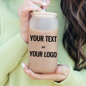 May include: A frosted glass tumbler with a bamboo lid and a clear straw. The tumbler is filled with a light brown liquid and features the text "YOUR TEXT or YOUR LOGO" in black. The tumbler is held by a person wearing a light green sweater.