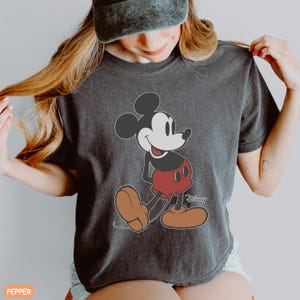 May include: Dark gray t-shirt featuring a classic Mickey Mouse graphic. The cartoon character is depicted in his iconic red shorts, brown shoes, and black ears. The Disney logo is visible on the shirt.