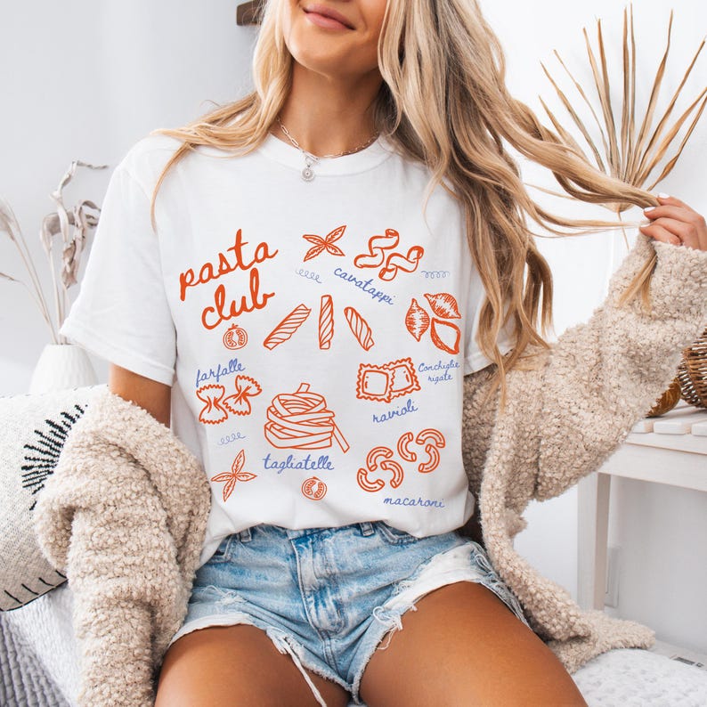 Pu&ograve; includere: T-shirt bianco con grafica di pasta rossa e blu. Le parole "pasta club" sono in alto, con illustrazioni di varie forme di pasta, tra cui farfalle, ravioli e tagliatelle. La maglietta &egrave; indossata con pantaloncini di jeans.