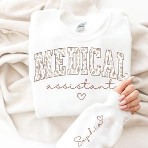 May include: Cream-coloured sweatshirt featuring the text "MEDICAL assistant" in a brown floral pattern. The name "Sophia" is written on the sleeve. A heart symbol is below the word "assistant" and next to the name "Sophia".