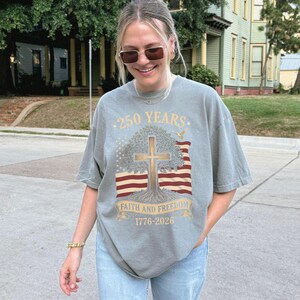 May include: A gray t-shirt with a graphic design commemorating 250 years. The design includes a cross, tree, American flag, and the text "Faith and Freedom 1776-2026". The shirt is a relaxed fit.