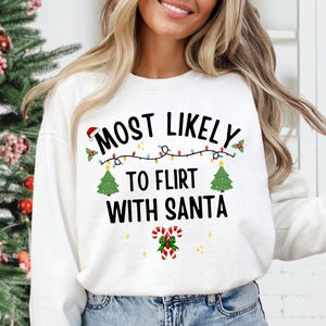 May include: A white long-sleeve sweatshirt featuring the black text MOST LIKELY TO FLIRT WITH SANTA. The design includes a string of colorful Christmas lights, two green Christmas trees, a small Santa hat, and two red and white candy canes.