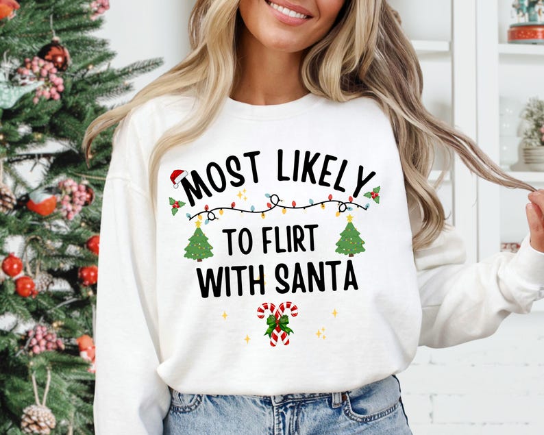 May include: A white long-sleeve sweatshirt featuring the black text MOST LIKELY TO FLIRT WITH SANTA. The design includes a string of colorful Christmas lights, two green Christmas trees, a small Santa hat, and two red and white candy canes.