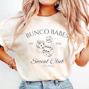 Peut inclure: Sweat-shirt crème avec l'inscription "Bunco Babes Social Club" et des graphiques de dés. Le design comprend l'année "2021" et la phrase "Let's Roll". Un vêtement décontracté.