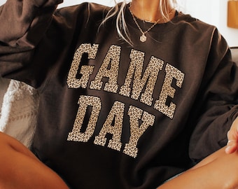 Può includere: Una felpa marrone scuro con la scritta "GAME DAY" a grandi lettere con stampa leopardata sul davanti. La felpa ha maniche lunghe e girocollo. Lo stile è casual.
