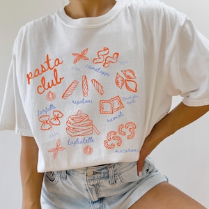 Pu&ograve; includere: T-shirt bianco con la scritta "pasta club" in arancione. La maglietta presenta illustrazioni arancioni di varie forme di pasta, tra cui farfalle, rigatoni e ravioli. La maglietta &egrave; abbinata a pantaloncini di jeans.