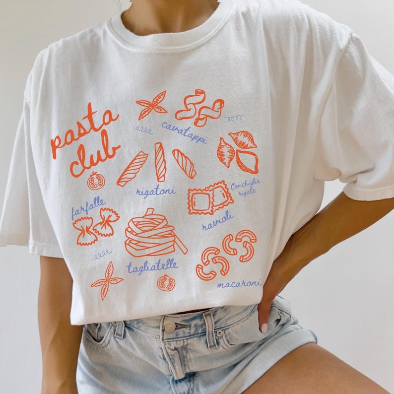 Pu&ograve; includere: T-shirt bianco con la scritta "pasta club" in arancione. La maglietta presenta illustrazioni arancioni di varie forme di pasta, tra cui farfalle, rigatoni e ravioli. La maglietta &egrave; abbinata a pantaloncini di jeans.