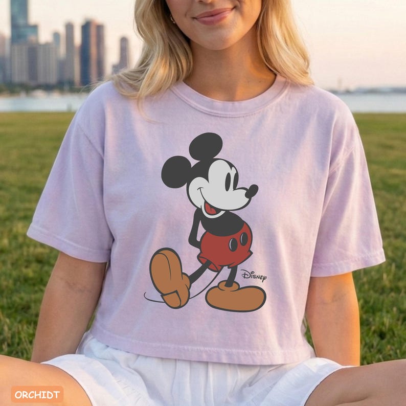 May include: A light purple t-shirt featuring a graphic of Mickey Mouse in a walking pose. Mickey Mouse is depicted with black ears, a white face, red shorts, and brown shoes. The Disney logo is visible. The word "ORCHIDT" is at the bottom left.