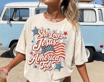 以下が含まれることがあります： She Loves Jesus and America Tooというテキストがプリントされたクリーム色の半袖Tシャツ。デザインには、なびくアメリカ国旗、赤と青の星、そして背景に色あせた青いストライプ模様が描かれています。