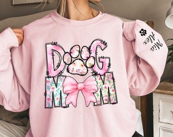 May include: A light pink sweatshirt with the words "Dog Mom" in a floral design. The paw print in the "O" is filled with a floral pattern. A pink bow is below the words. The sleeve has a paw print and the names "Mia" and "Alex".