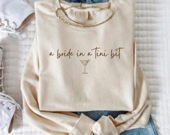 May include: Cream-colored sweatshirt with the phrase "a bride in a tini bit" in brown script, along with a martini glass illustration. A gold chain necklace rests on the sweatshirt. The sweatshirt is paired with blue jeans and white sneakers.
