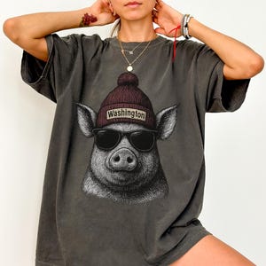 May include: Dark gray t-shirt featuring a pig wearing sunglasses and a maroon beanie with "Washington" printed on it. The pig graphic is a detailed illustration. The shirt has short sleeves and a relaxed fit.