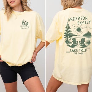 May include: A pale yellow short-sleeve t-shirt featuring a dark green graphic of two chairs by a lake with pine trees and the sun. The front chest displays a small version of the design, while the back features the text Anderson Family Lake Trip Est. 2026.