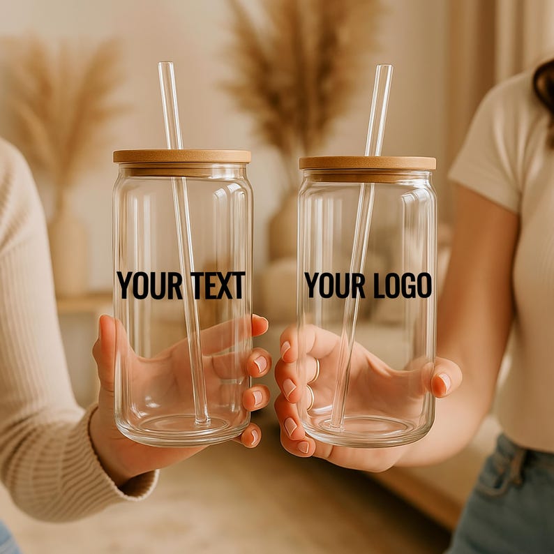 May include: Two clear glass beverage cups with bamboo lids and clear straws are held up. The text "YOUR TEXT" and "YOUR LOGO" are printed on the cups. The background is blurred, with neutral tones.