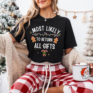 May include: A black short-sleeved t-shirt featuring the white text MOST LIKELY TO RETURN ALL GIFTS, decorated with colorful string lights, two gingerbread cookies, and two candy canes with green holly leaves and red berries.
