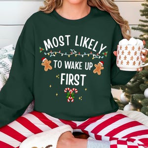 May include: A dark green crewneck sweatshirt featuring the white text "MOST LIKELY TO WAKE UP FIRST" with graphics of two gingerbread people wearing Santa hats, a string of colorful Christmas lights, and two candy canes with holly leaves.