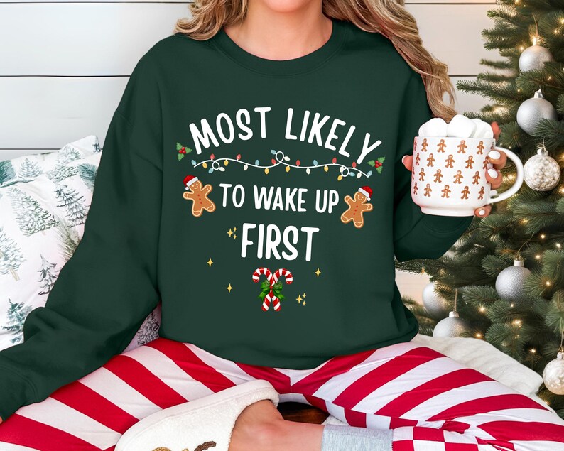 May include: A dark green crewneck sweatshirt featuring the white text "MOST LIKELY TO WAKE UP FIRST" with graphics of two gingerbread people wearing Santa hats, a string of colorful Christmas lights, and two candy canes with holly leaves.