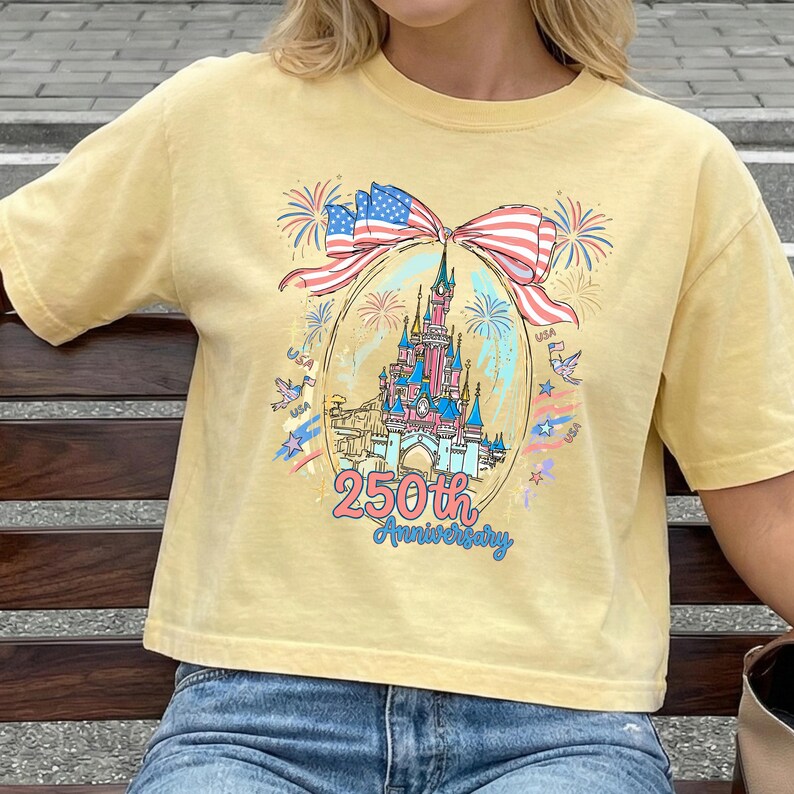 K&ouml;nnte beinhalten: Hellgelbes T-Shirt mit einer Grafik eines Schlosses, Feuerwerks und einer Schleife in Rot, Wei&szlig; und Blau. Der Text "250th Anniversary" ist in blauer Schrift. Das T-Shirt hat eine hellgelbe Farbe.